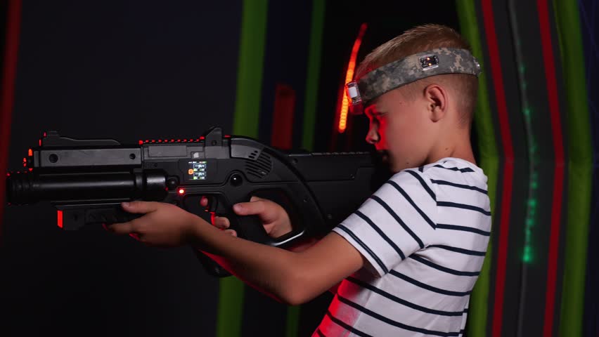 Close-up of an active boy playing laser tag with friends, he holds a laser weapon and aims at the camera. A game arena with bright stripes on the wall. An active play area for children.