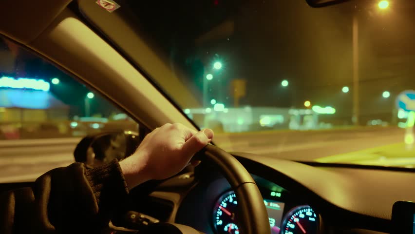 Hand gripping steering wheel while unrecognizable man driving past gas station at night. Male steering car, holding wheel while passing fuel station under lights. Driving through nighttime road