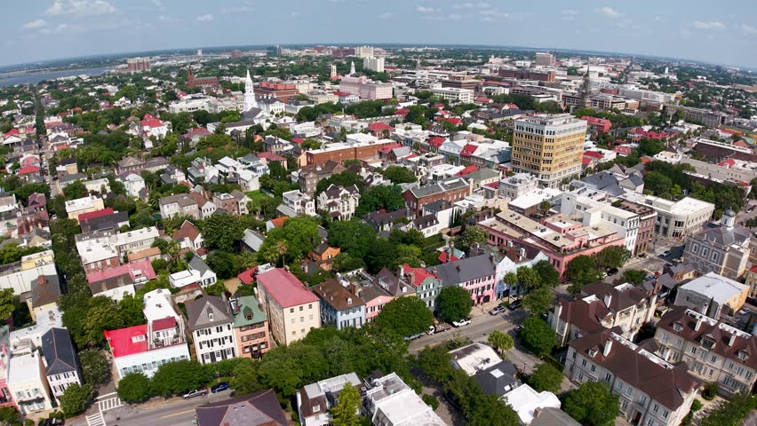 Aerial drone view of Charleston, South Carolina cityscape with colorful historic buildings, green trees and a bird flying across the skyline on a sunny day.