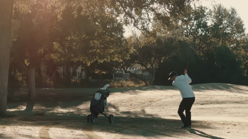 Golfer with a walking cart beside him hits a fairway wood in slow motion, carving a clean divot as the ball launches down the fairway toward camera