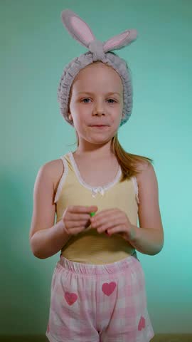 Young girl with fuzzy bunny ears headband enjoying a long, blowing a children's whistle for a birthday party. Child in heart-patterned pink shorts and yellow tank top laughing.