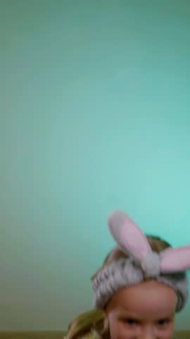 Young girl with fuzzy bunny ears headband enjoying a long, blowing a children's whistle for a birthday party. Child in heart-patterned pink shorts and yellow tank top laughing.