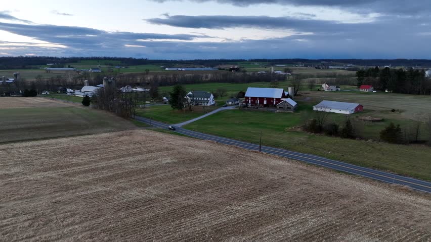Rural American farmland at dusk with red barn, white farmhouses, open fields and quiet country road cutting through rolling landscape under cloudy evening sky. Aerial flyover. Virginia, USA.