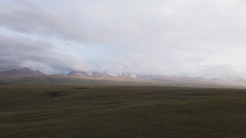 Kyrgyzstan - Soft Light Falls Over a Wide, Empty Plain as Distant Snow-tipped Mountains Fade Beneath Heavy Clouds - Aerial Drone Shot