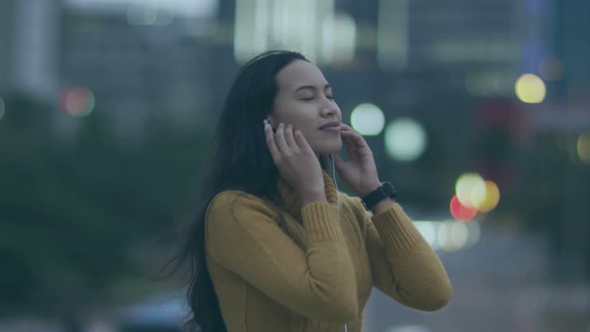 Woman plugging earphones, hearing music, opening arms amid bokeh, promo THANK YOU overlay above her. Urban, outdoor, dusk, headphones, citylights, outstretched, serene
