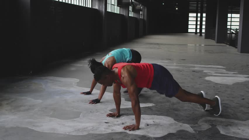Pair starting crouch, doing fast jumps-into pushups for fitness, 3D GOOD VIBES overlay over pair. Warehouse, industrial, athletes, dynamic, teamwork, sidelight, concrete