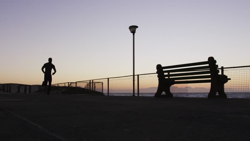 Male jogger starting run on sea, camera tightening while text overlays obscuring runner for fitness. Silhouette, coastal, promenade, dawn, bench, seagull, motion