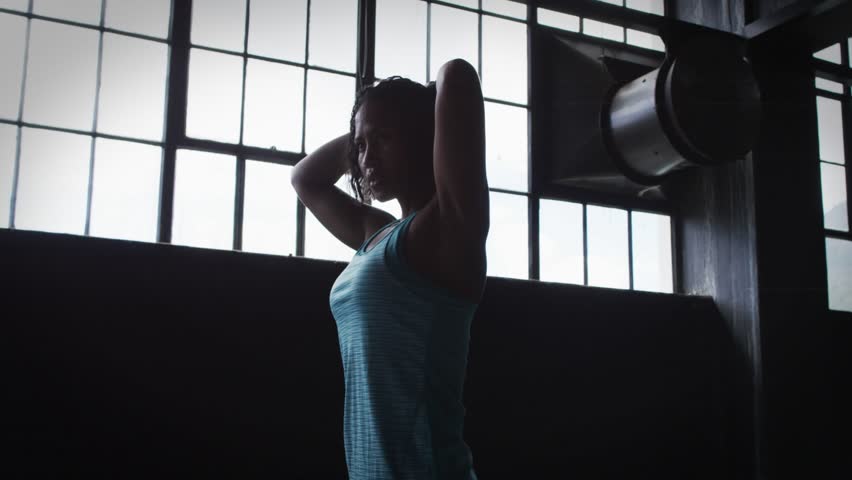 Woman stretching and adjusting hair in gym loft, glitching GOOD VIBES graphic overlaying silhouette. Backlit, industrial, windowpane, female, fitness, stretch, posture