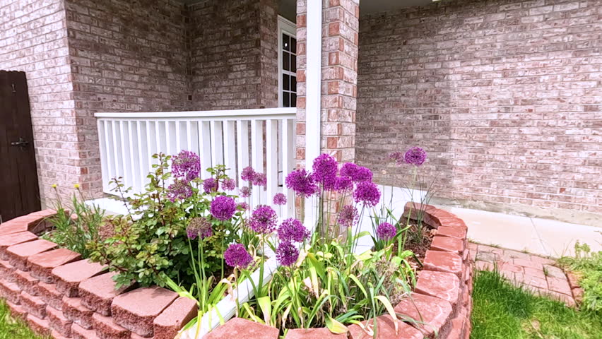 The front porch of a charming brick house featuring a white door and a well-maintained garden bed with blooming purple flowers. The neatly trimmed lawn and inviting entryway enhance the homes curb