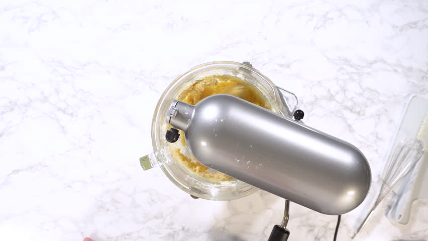Overhead shot of stand mixer blending pale cookie dough mixture in glass bowl on marble countertop.