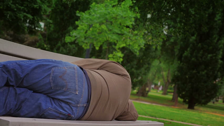 A homeless man sleeps alone on a park bench.