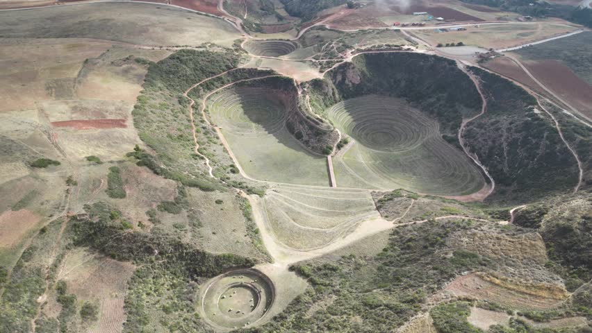 Drone view of Moray ruins in Peru, showcasing circular Incan terraces set into the Andes mountains under sunny skies with scattered clouds.