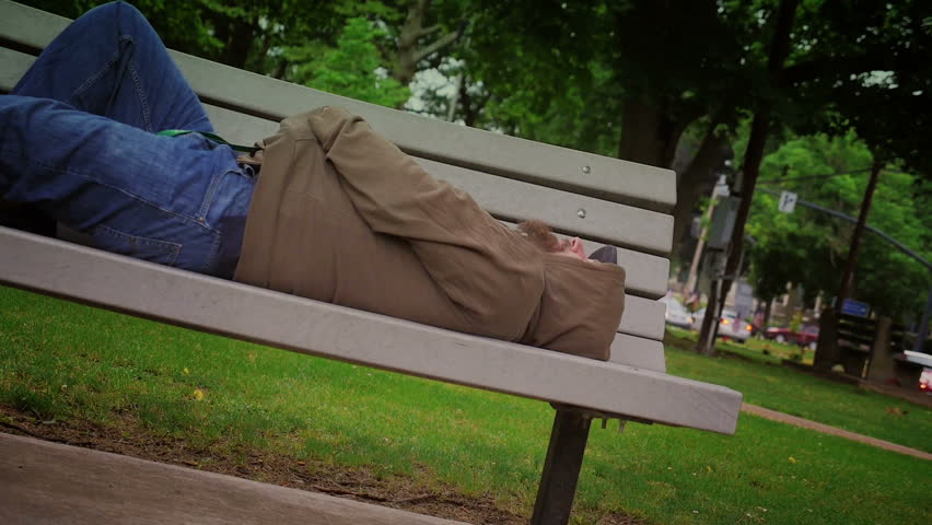 A homeless man sleeps alone on a park bench.