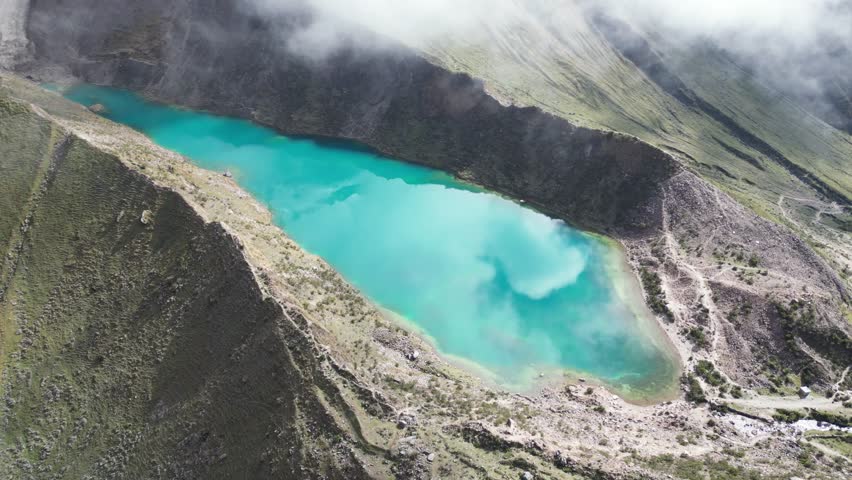 Aerial drone shot of Humantay Lake, Peru, where turquoise glacial water mirrors bold cloud formations in clear sunlight.