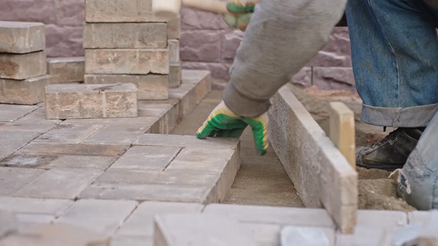 Worker laying paving stones and using a mallet, performing manual labor on a construction site outdoors.