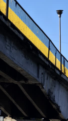Low angle view of an old concrete bridge with its railing painted in the national colors of Ukraine, blue and yellow, standing as a symbol of support and solidarity against a clear blue sky
