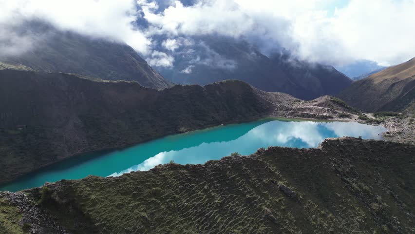 Aerial view of Humantay glacial lake in the Andes, showing turquoise water reflecting dramatic cloud patterns in clear, sunlit conditions.