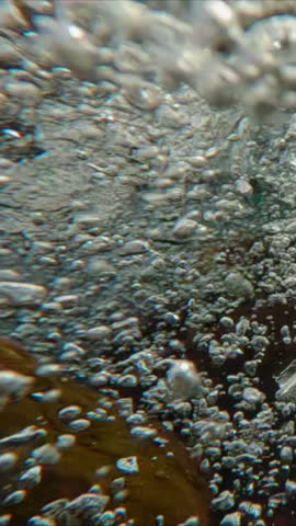 Vertical footage, Underwater view of many air bubbles in a turbulent mountain stream flowing over a rocky bottom with algae, macro video, super slow motion