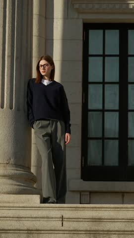 Long lens parallax shot of stylish young woman in glasses and loose trousers posing against massive stone column on Seokjojeon palace steps at golden hour