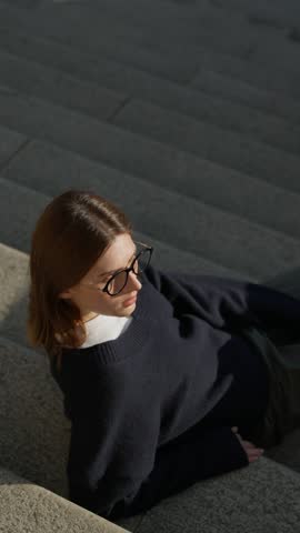 Stylish slim young woman in oversized navy sweater and round glasses leans back on wide stone steps, posing in warm sunset light - vertical slow motion, parallax shot
