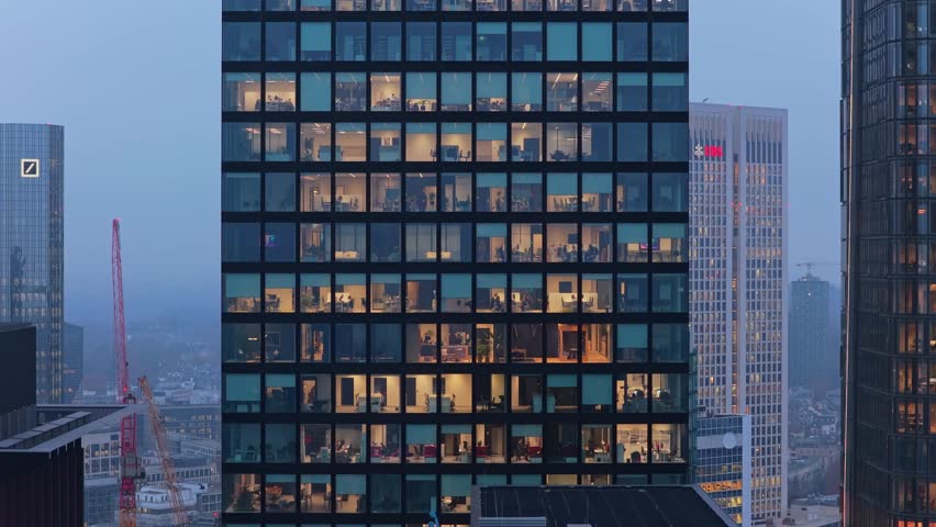 Aerial view of a glass skyscraper at dusk with office workers visible inside their illuminated workspaces in Frankfurt's financial district