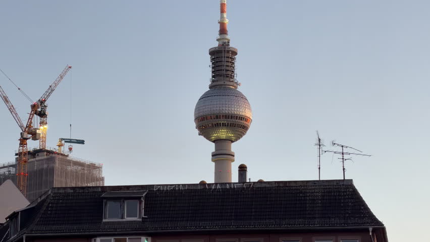 Handheld shot of Berlin TV Tower, Fernsehturm, rising above city buildings and cranes in Alexanderplatz at sunset, symbolizing urban development