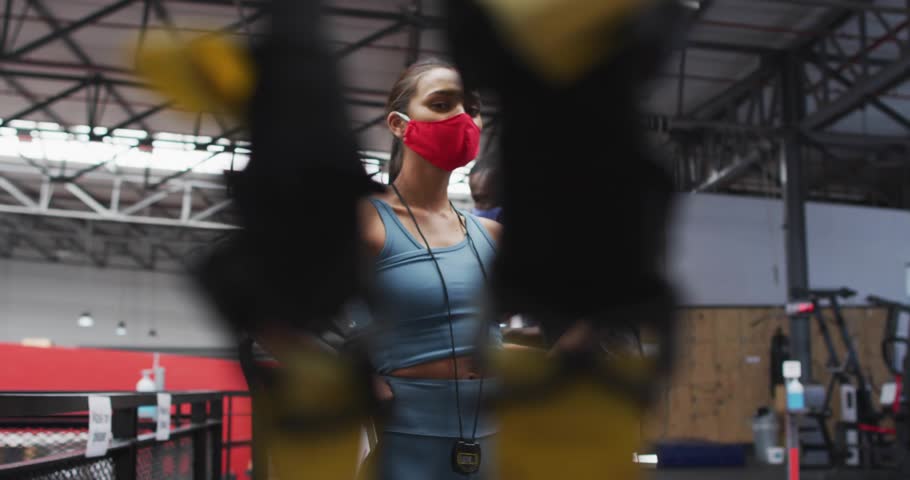 Woman hands-on-hips coaching, gym straps shifting revealing her while man rowing, virus overlaying. Fitness, training, partners, instructor, cardio, rower, trx