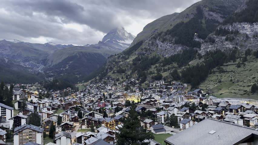 Zermatt village stretches across the alpine valley beneath the Matterhorn, surrounded by forested hills, rugged peaks, and peaceful evening light illuminating classic Swiss mountain architecture.