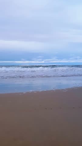 Low angle vertical shot of stormy coastal surf water waves rolling on sand shallow of empty beach on Baltic sea shore under blue cloudy sky at overcast day. Desolate seascape natural background.