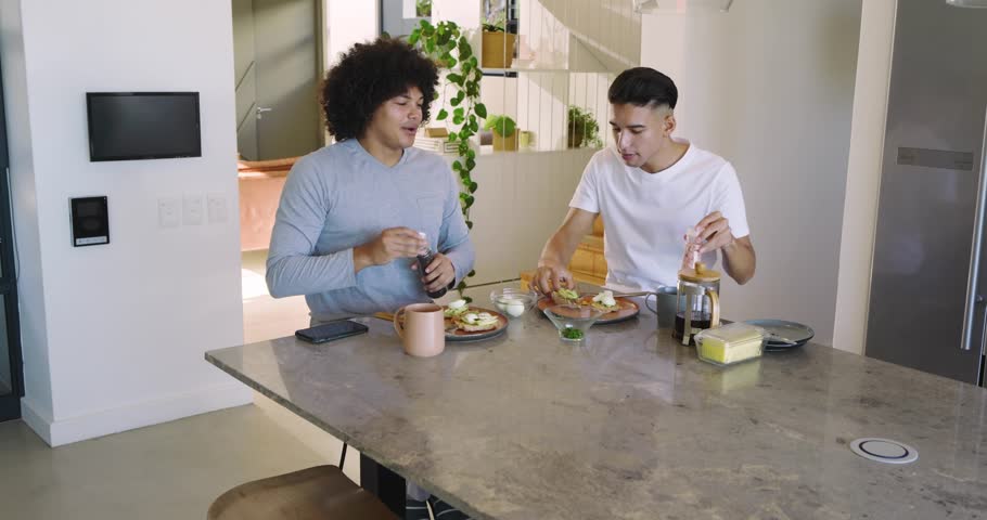 Two men sitting island eating, gold confetti falling and causing feeding, celebrating lifestyle. Friends, breakfast, kitchen, coffee, candid, festive, laughter