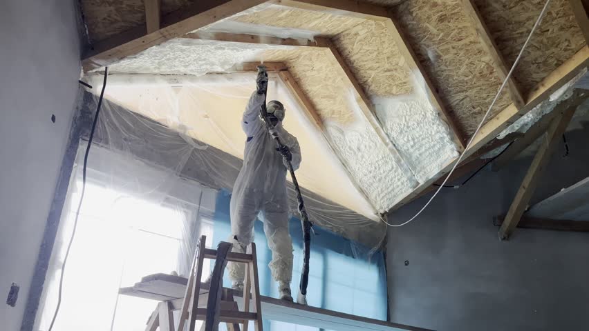 Professional construction worker wearing a protective suit and mask spraying polyurethane foam to insulate the wooden roof structure of a house under construction for thermal and soundproofing