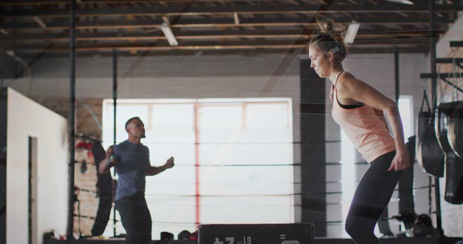 Timer starting, woman doing plyo-box jumps for fitness, man jump-roping, blue swirls accenting. Plyometric, training, warehouse, industrial, partner, movement, dynamic