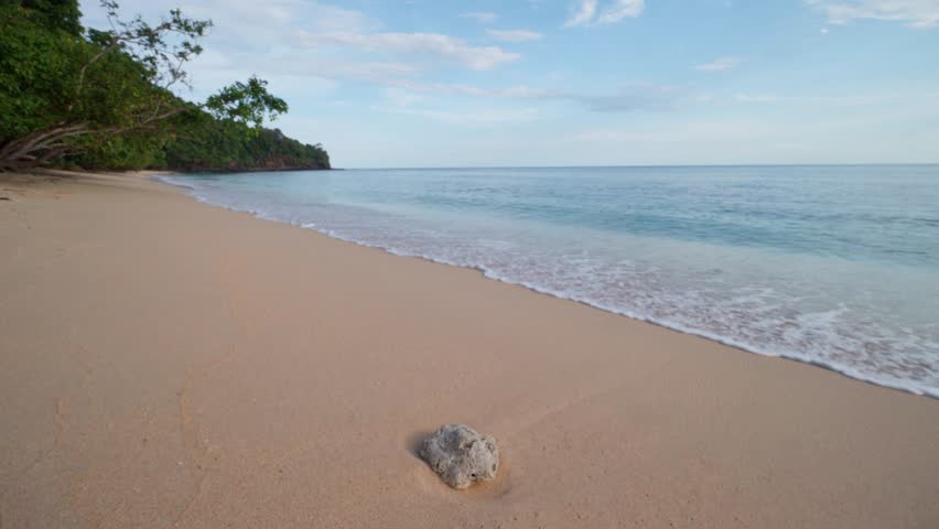 A wide, serene tropical beach with fine sand, gentle wave, and clear blue sky, featuring one rock in the foreground. Peaceful nature background.
