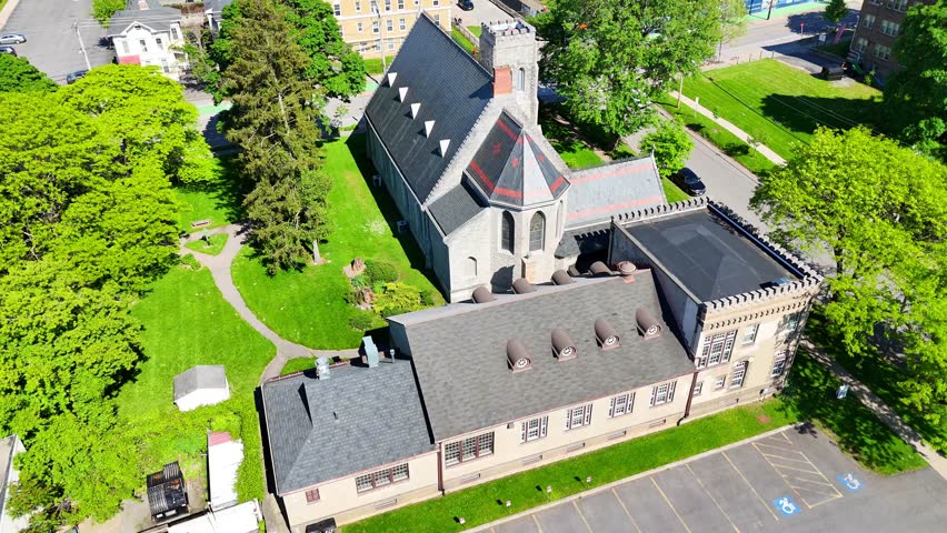 Aerially circling an Episcopalian church designed in the architectural style of gothic revival surrounded by the counterparts of a city.