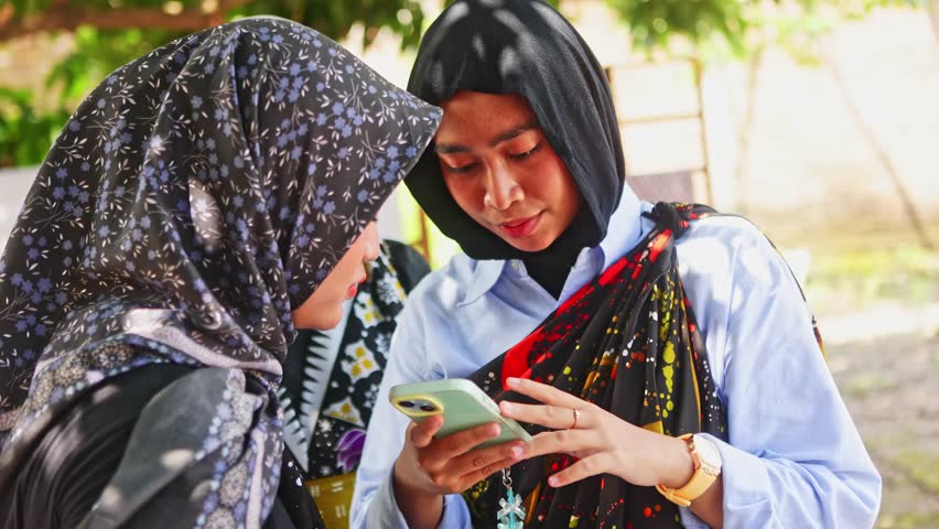 Beautiful indonesian women wearing traditional batik and hijabs smiling and laughing together while looking at a smartphone screen, showcasing cultural heritage and modern connection