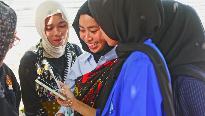 Group of young indonesian women wearing traditional batik and modern hijab looking at a smartphone, smiling and laughing together while sharing content with genuine happiness and connection