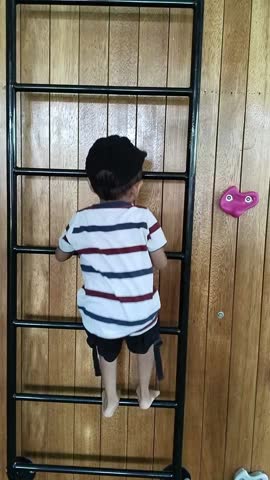 Asian boy wearing a black hat focused on climbing a black iron ladder attached to a wooden wall to practice his gross motor skills, causing his hat to fall