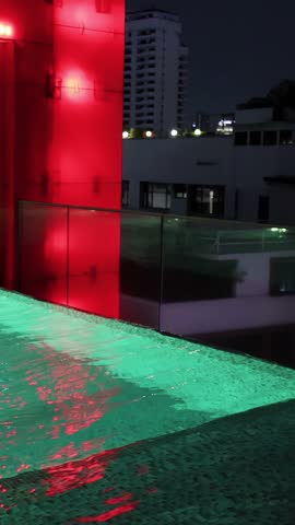 Camera pans across illuminated rooftop swimming pool, neon red columns, city skyline, and night reflections