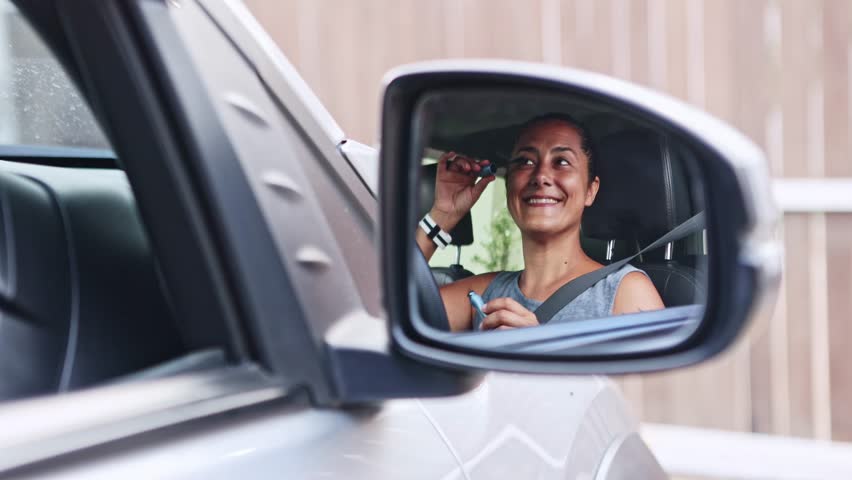 Reflection of a smiling woman in a car side mirror applying mascara to her eyelashes, getting ready for a business meeting or a date while sitting in her vehicle with her seatbelt fastened