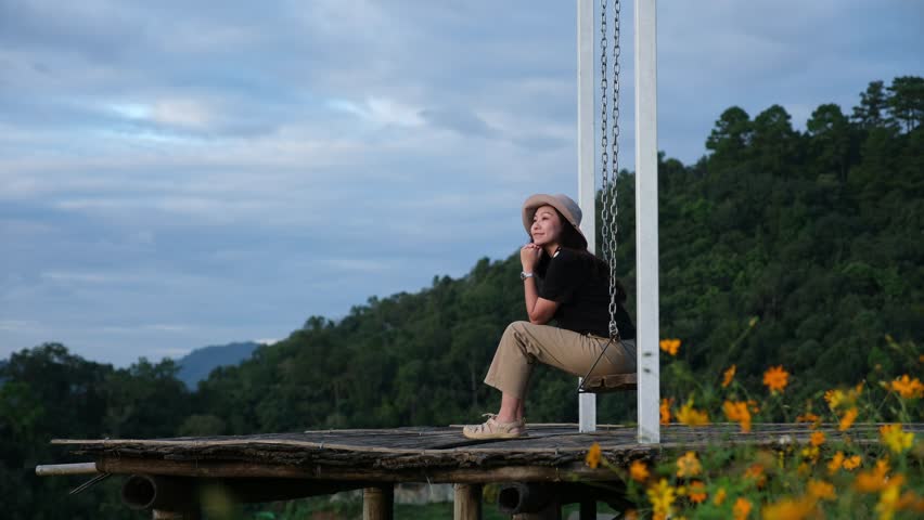 A woman sitting on a swing with a beautiful flower garden and mountain on cloudy day