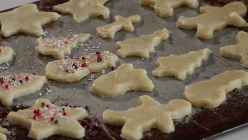 A close up of red and green sprinkles being dropped a variety of holiday shaped cookies on a cookie tray.