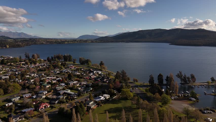 Te Anau Town in New Zealand’s South Island. lakeshore, marina area, residential neighborhoods, surrounding greenery and Fiordland mountains under bright afternoon light. Aerial view.