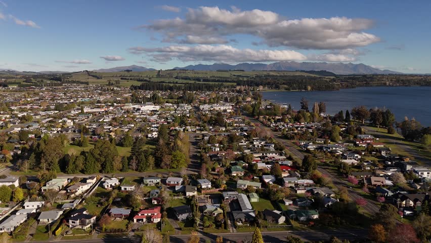 Aerial wide shot of Te Anau in New Zealand’s South Island, showing lakeside town, residential areas, greenery and Fiordland mountains under bright daylight with scattered clouds. Peaceful neighborhood