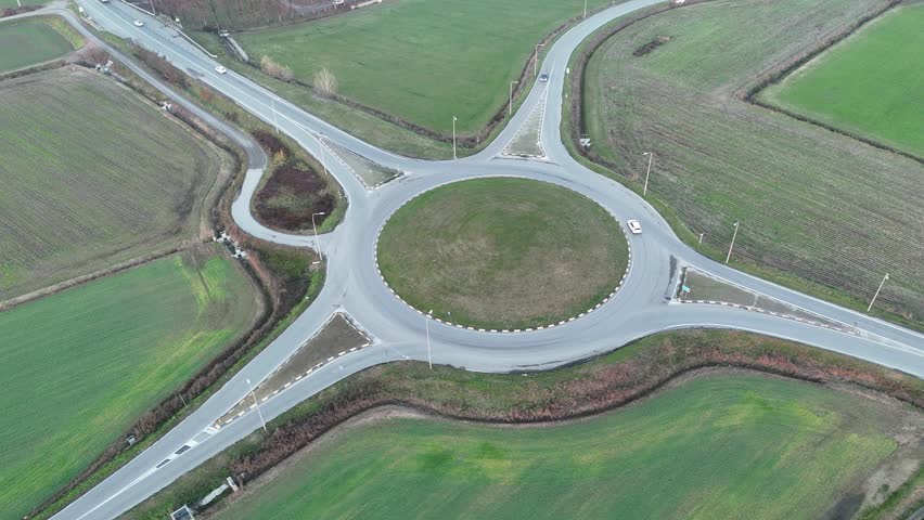 Contemporary roundabout in Strada Paullese SP415 road, linking rural roads in Lombardy, cars and trucks moving through green and earthy farmland under golden hour light and a orange sunset sky