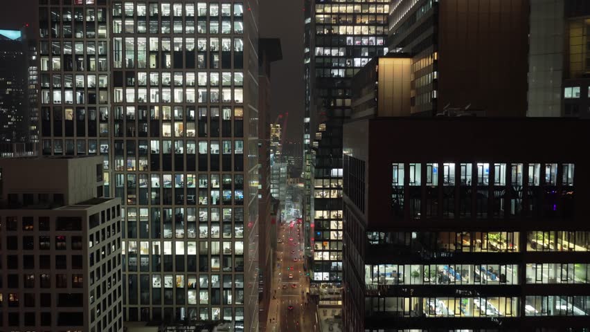 Aerial view moving between skyscrapers with lit windows, showing people working late in their offices. Financial district of a Western Europe metropolis