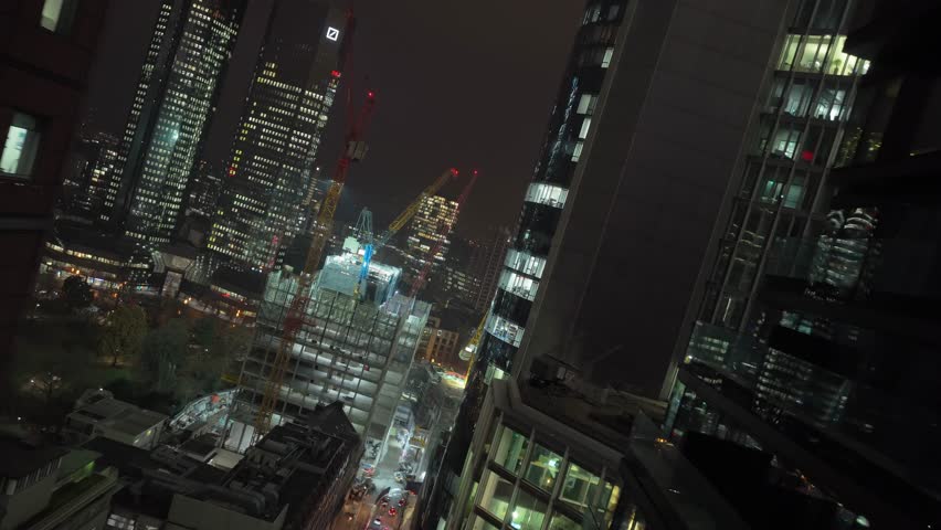 Aerial view flying downwards between towering skyscrapers at night. Luminous office windows reveal bustling financial district