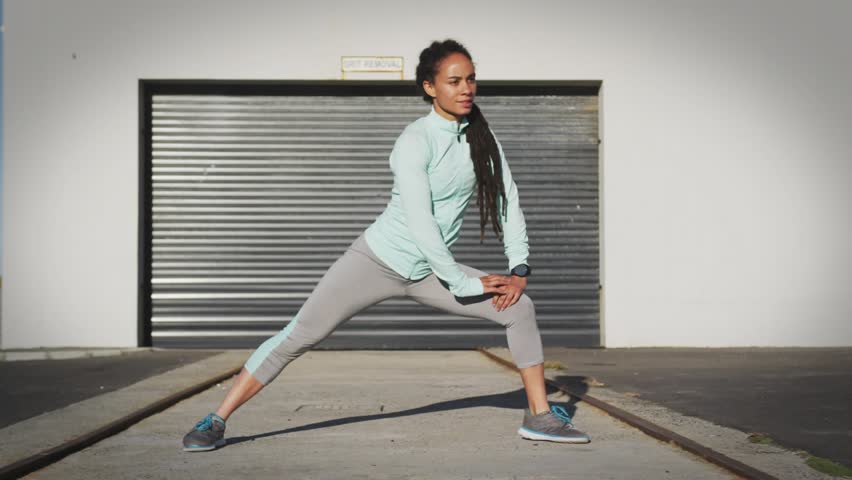Woman warming up, lunging laterally while GOOD VIBES text glitching, settling in front for fitness. Urban, industrial, exterior, stretch, workout, trainer, sunlit
