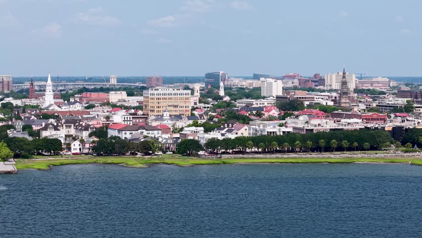 Aerial drone shot over the waterfront city skyline of Charleston, South Carolina, showing historic buildings, church steeples, and harbour waters on a sunny day.