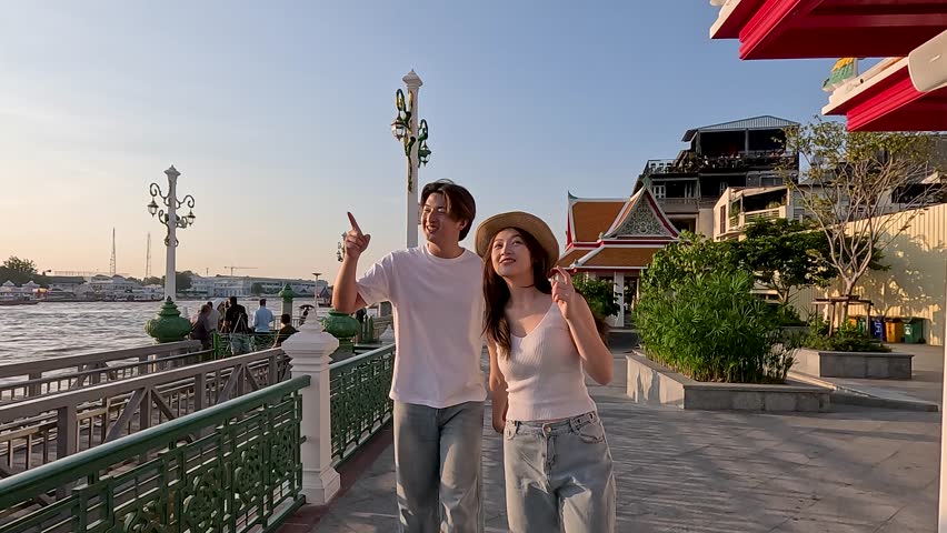 Smiling young couple strolls along riverside promenade at golden hour with smooth tracking shot