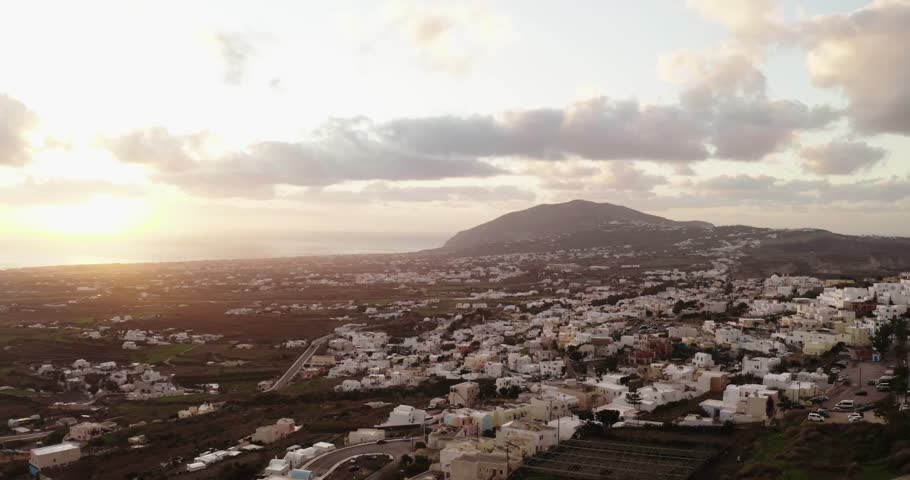 Drone view over a sprawling white Cycladic town on the flat part of Santorini. The sunset shines dramatically through clouds, illuminating the town and a large volcanic mountain in the distance.